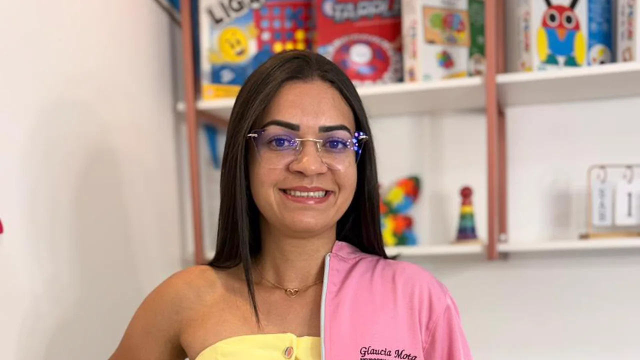 Glaucia Amanda sorrindo, com jaleco rosa sobre roupa amarela, em frente a uma estante de jogos