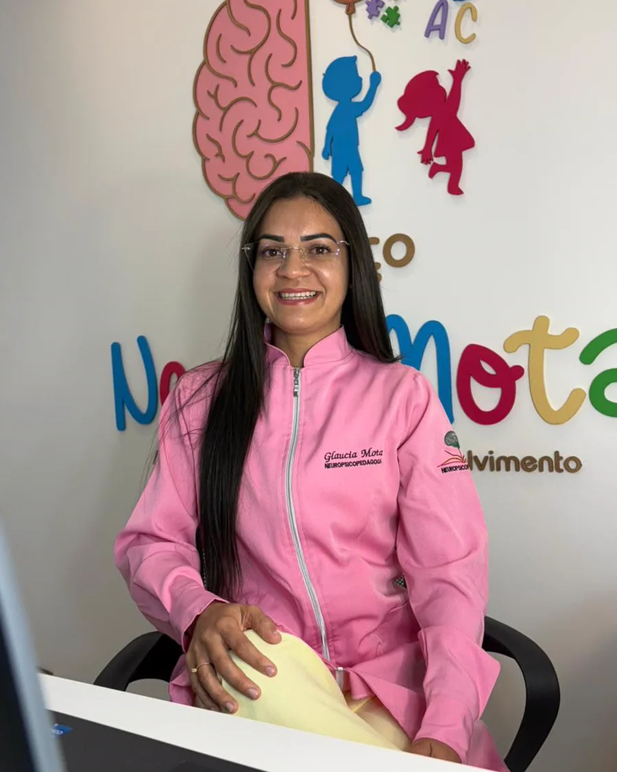 Retrato de Glaucia Amanda sentada à mesa, de jaleco rosa, com o painel do Espaço Neuromota ao fundo